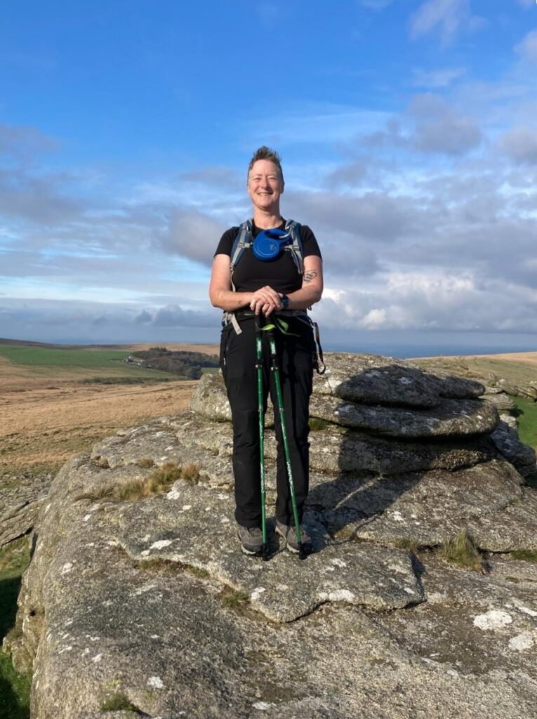 Zoe standing on a Dartmoor tor with hiking backpack, green rolling moorland and ocean visible in background